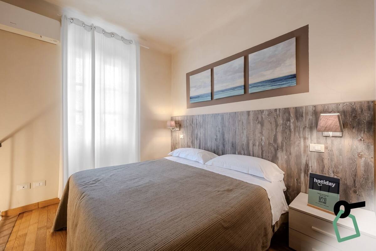 A hotel room with a bed, a wooden headboard, a nightstand, a wall-mounted lamp, and a painting of the sea.
