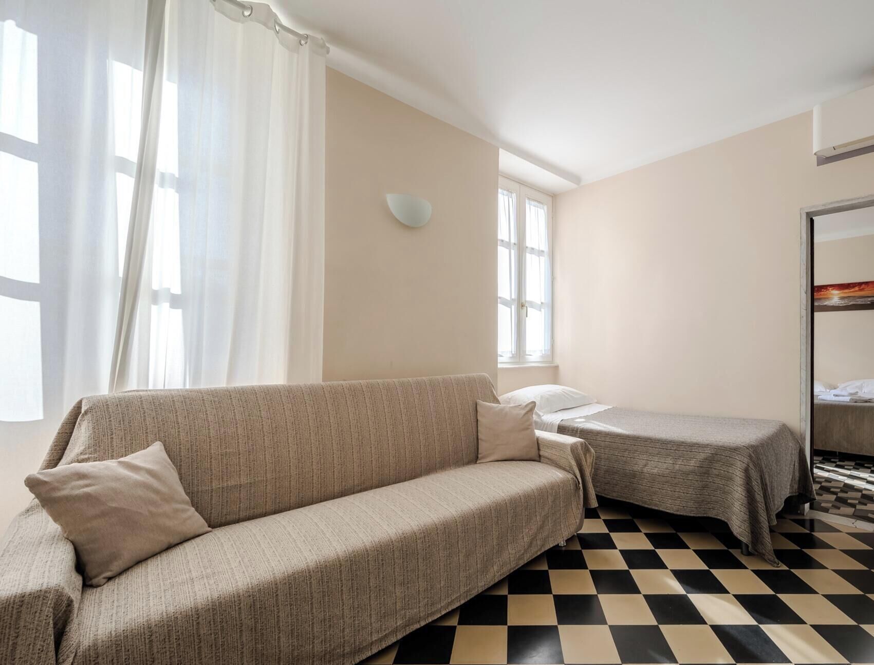 A room with a sofa, a bed, a checkered floor, and a window with curtains.