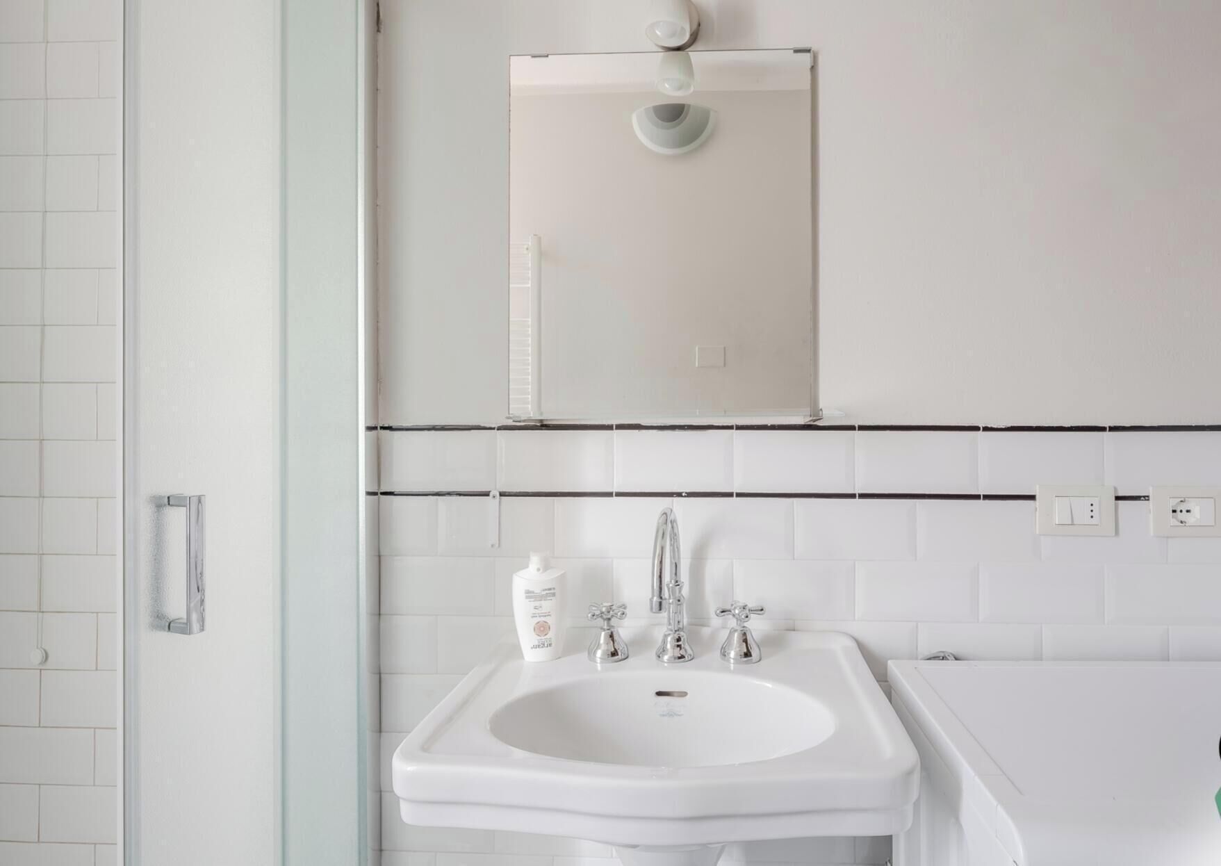 Een badkamer met een wit wastafelmeubel, twee kraanwerkstukken, een spiegel en een lamp boven de spiegel.