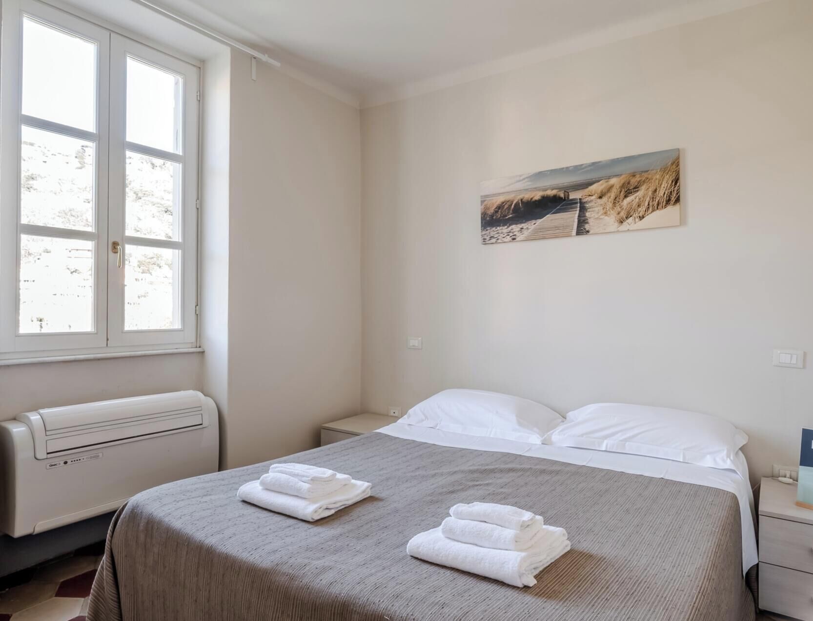 A hotel room with a bed, a window, a radiator, and a painting on the wall.