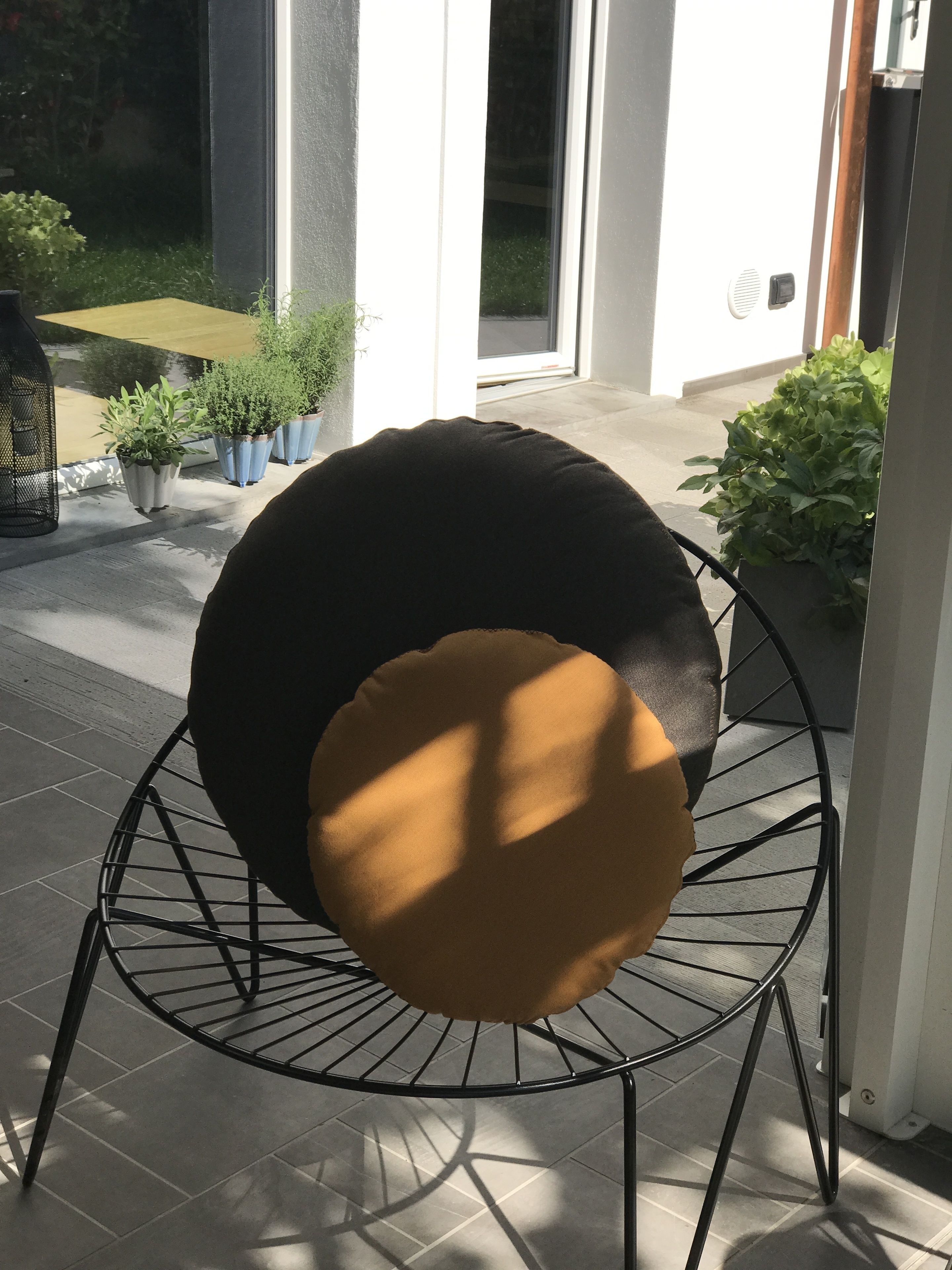 A round, wire-framed chair with a black and brown cushion, positioned on a tiled patio with potted plants and a door in the background.