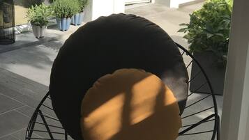 A round, wire-framed chair with a black and brown cushion, positioned on a tiled patio with potted plants and a door in the background.
