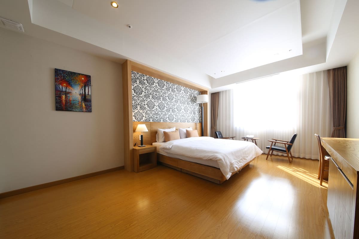 A modern hotel room with a wooden floor, a large bed, a desk, and a chair. There is a painting on the wall and a window with curtains.