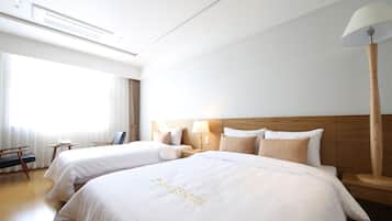 Habitación de hotel con dos camas, cabecero de madera, mesita de noche con lámpara y ventana con cortinas.