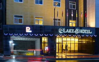 Entrada de um hotel com placa de estacionamento, várias bandeiras e fachada bem iluminada.