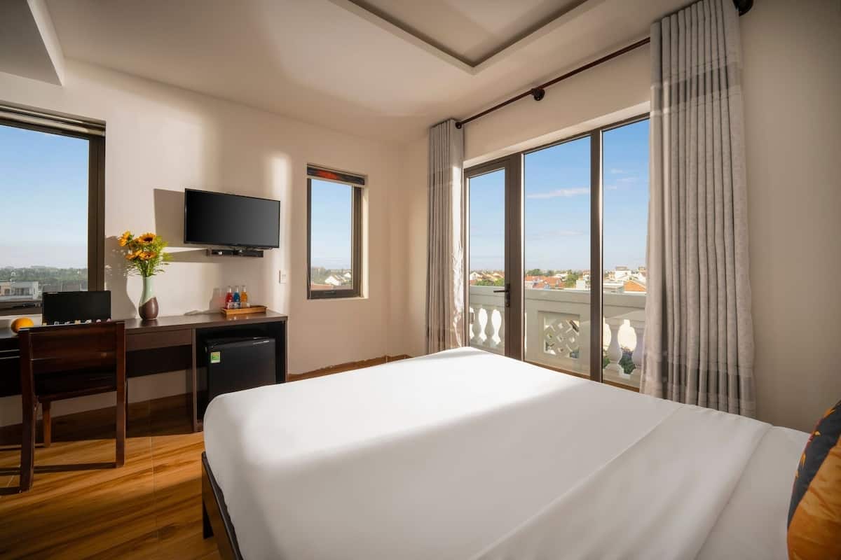 A hotel room with a bed, a desk with a computer, a TV, and a view of the city.