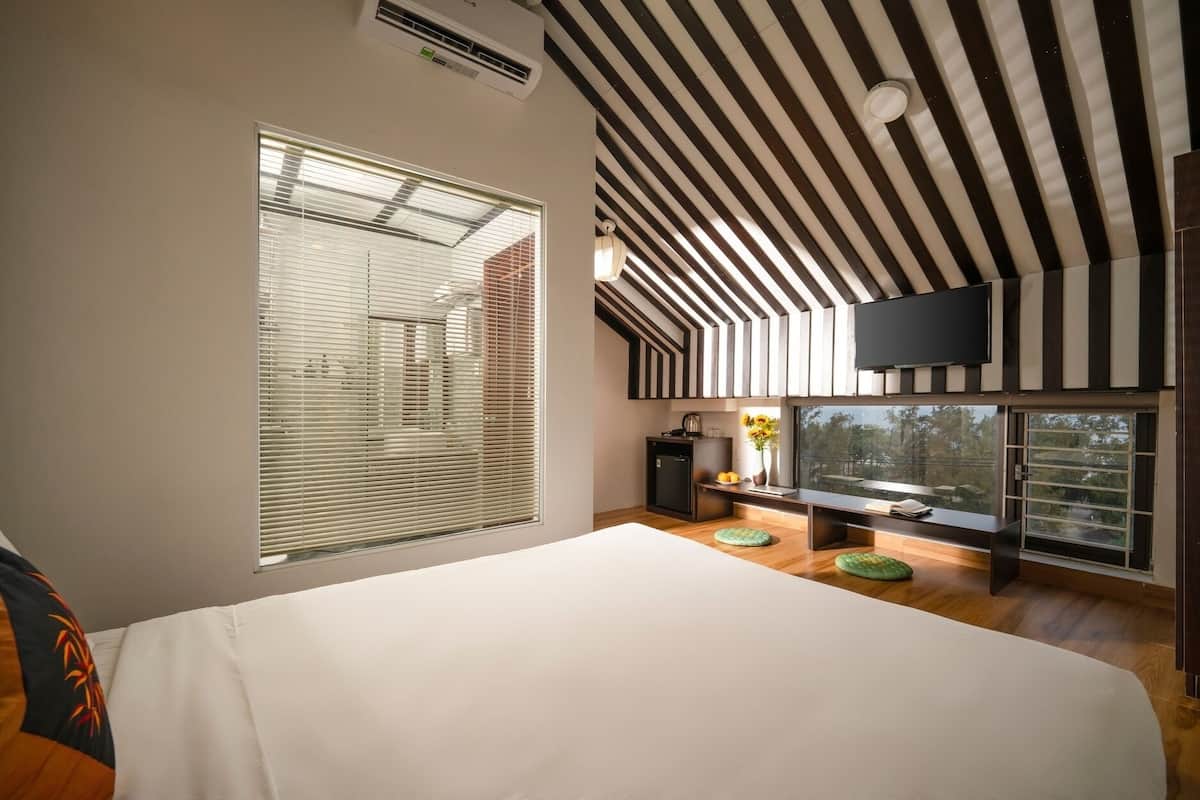 A modern bedroom with a large bed, a TV mounted on the wall, and a window with blinds.