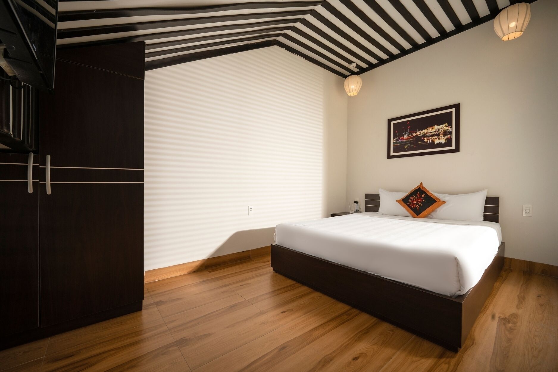 A modern bedroom with a large bed, wooden flooring, a sloped ceiling with black beams, and a cityscape painting on the wall.