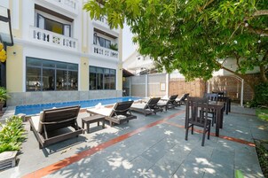 Zona de piscina con sillones de descanso y una pequeña mesa, rodeada por un edificio con grandes ventanales y un muro de ladrillo.