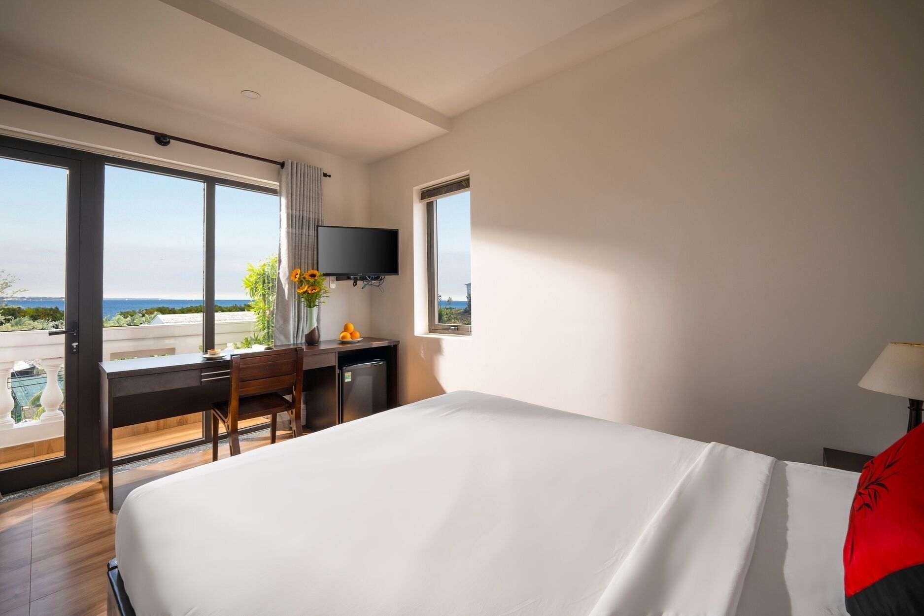 A hotel room with a bed, a desk with a TV, a window with a view of the sea, and a balcony.