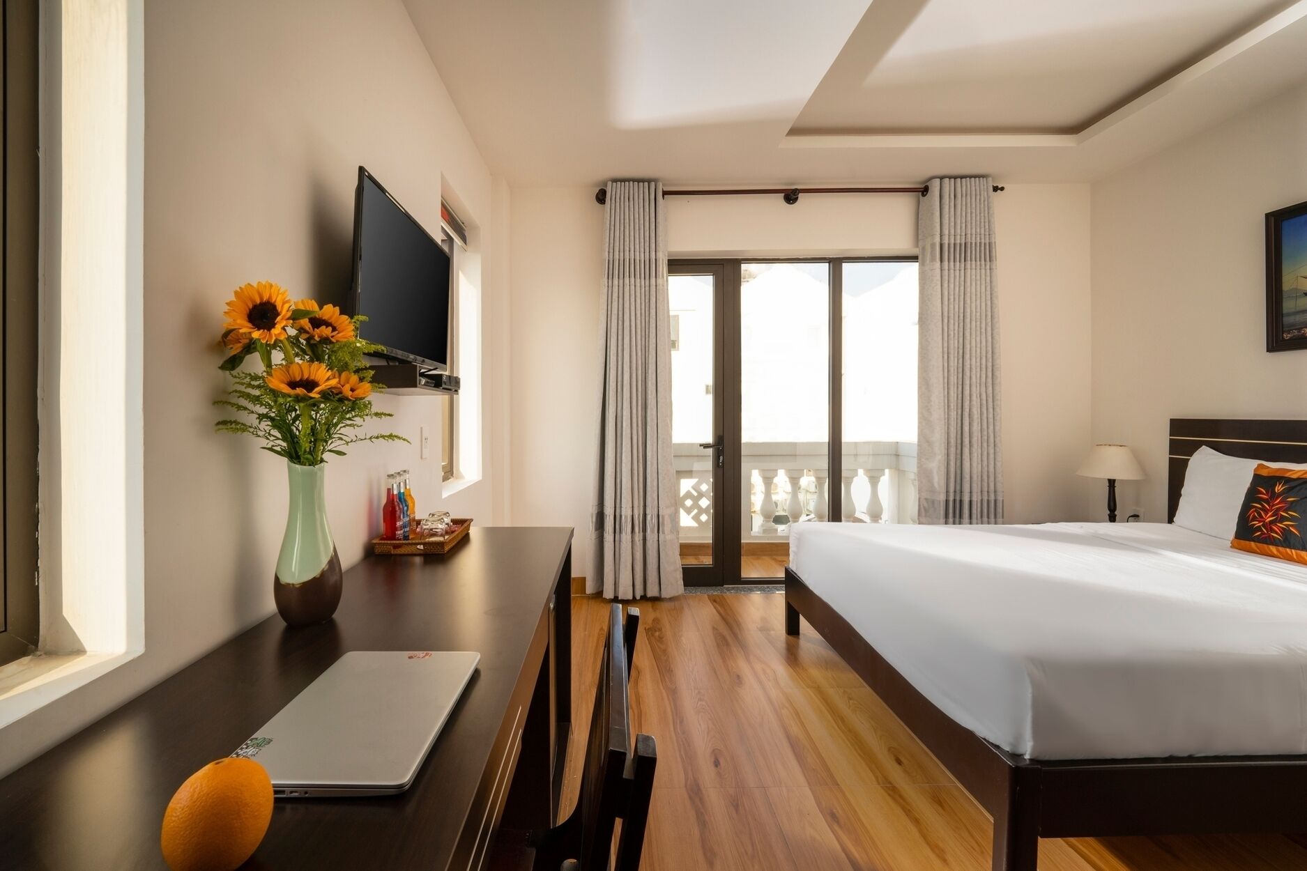 A hotel room with a bed, a desk with a laptop, a vase with flowers, a TV, and a balcony with curtains.