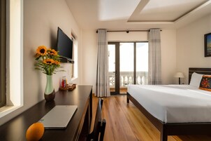 Habitación de hotel con cama, escritorio con una computadora portátil, florero con flores, televisor y balcón con cortinas.