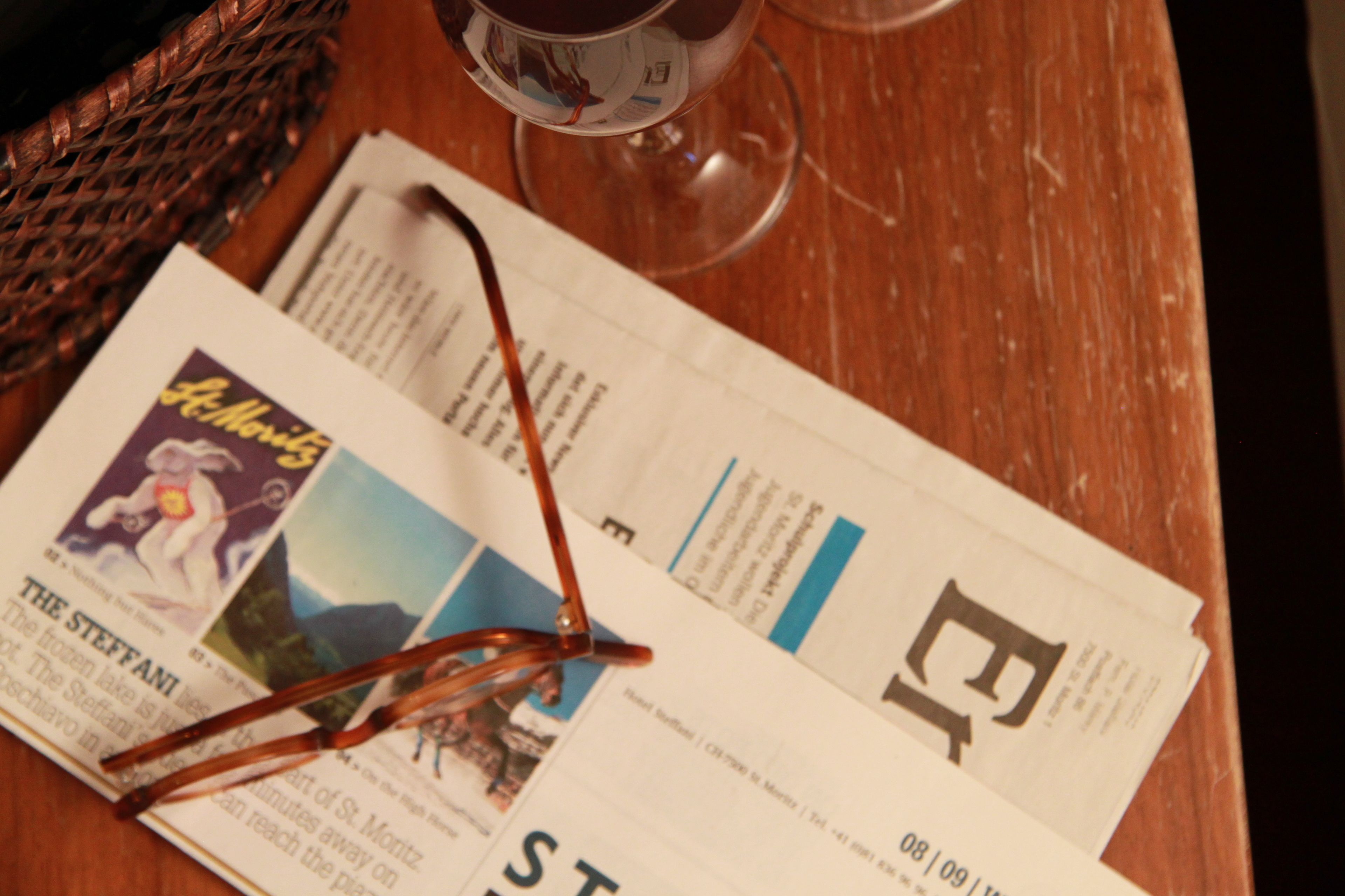 Un periódico, un par de gafas y una copa de vino tinto sobre una mesa de madera.