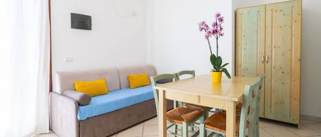 Een ruime, goed verlichte kamer met een bank, een eettafel met stoelen, een potorkidee en een houten garderobe.