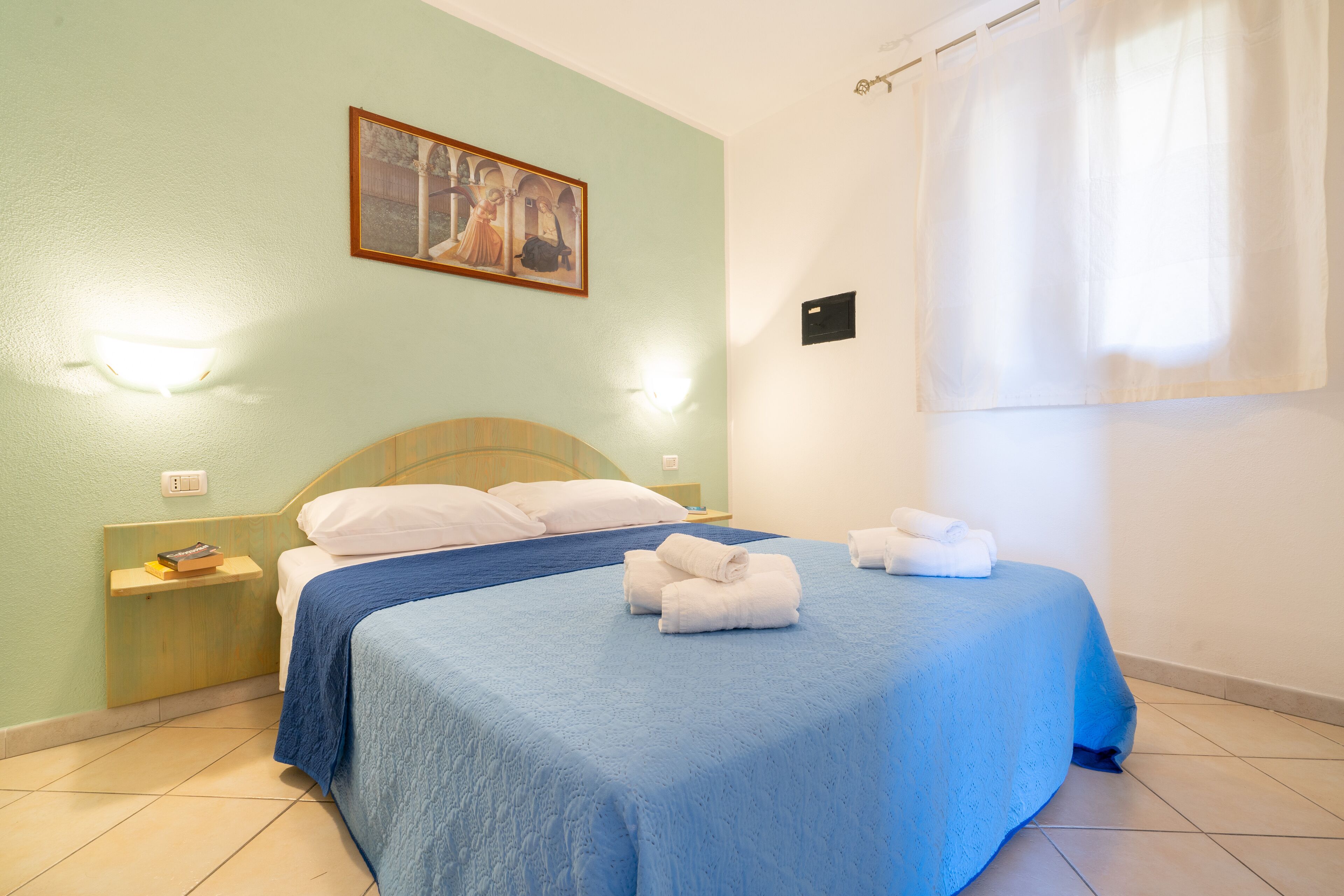Un dormitorio con una cama, una mesita de noche, una lámpara de pared, un cuadro y una ventana con cortinas.