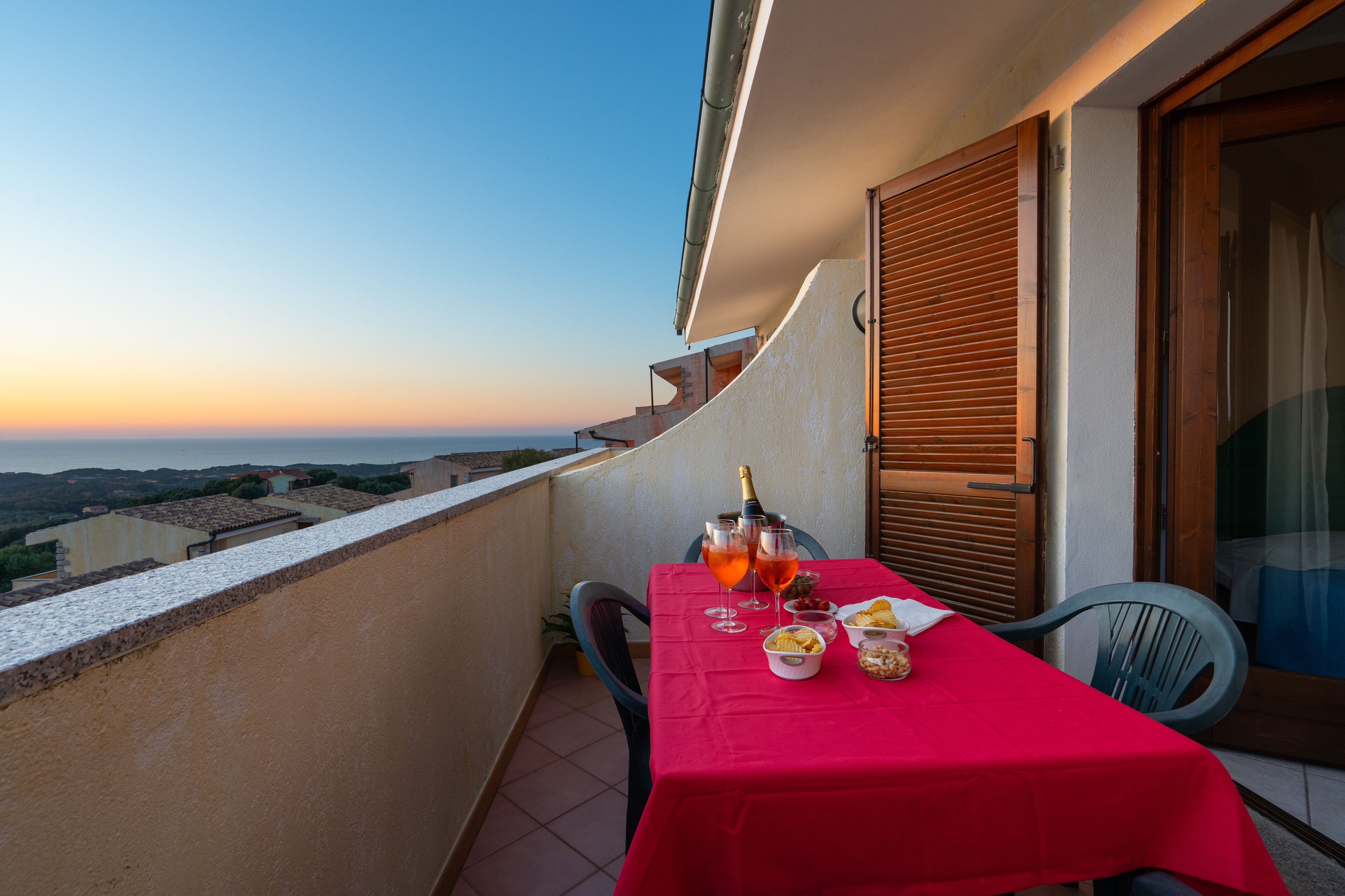A balcony with a red table set for two, a bottle of champagne, and snacks.