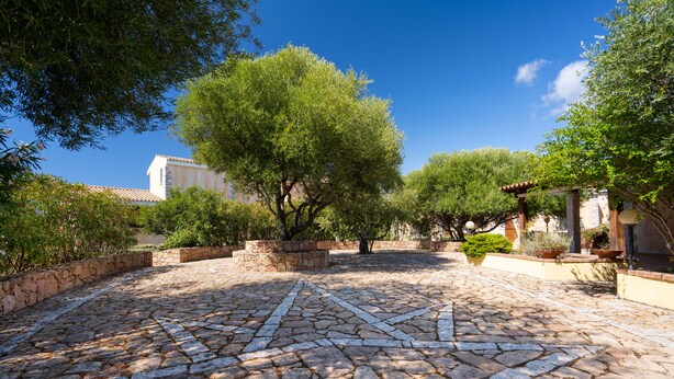 A courtyard with a cobblestone floor, surrounded by trees and a building with a tiled roof. - Residence Costa del Turchese by PendraHolidays (Badesi)