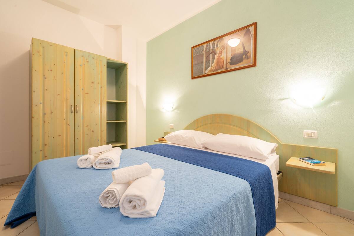 Ein ordentlich bezogenes Bett mit blauer Bettwäsche und weißen Kissen, ein Holzschrank mit Spiegel sowie ein kleiner Nachttisch mit Buch und Lampe.