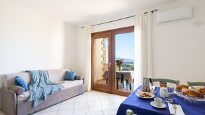 A room with a sofa, a dining table with breakfast items, and a view through a glass door. - Residence Costa del Turchese by PendraHolidays (Badesi)