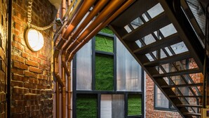 Eine moderne Treppe mit Metallgeländern und freiliegenden Rohren, die zu einem Balkon mit Blick auf eine Ziegelwand und grünes Kunstgrün führt.