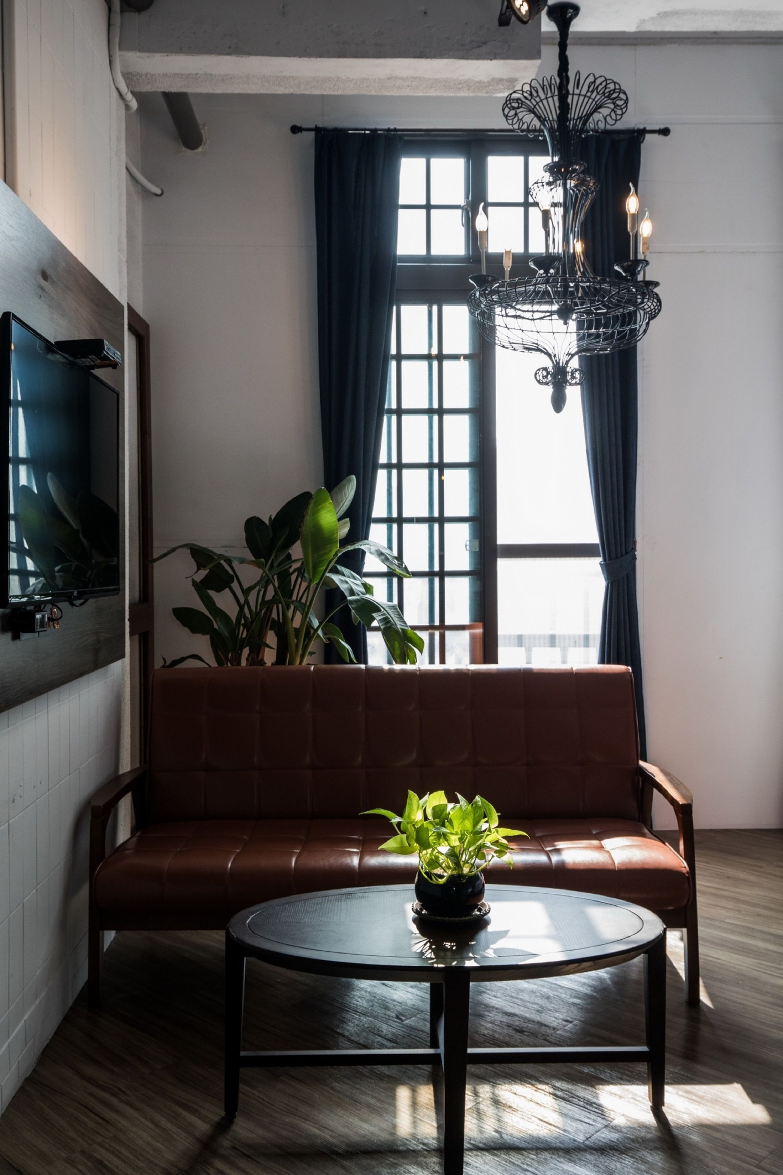 Ein Zimmer mit einem braunen Ledersofa, einem runden Tisch mit einer Pflanze, einer Kristallleuchtre und einem an der Wand befestigten Fernseher.