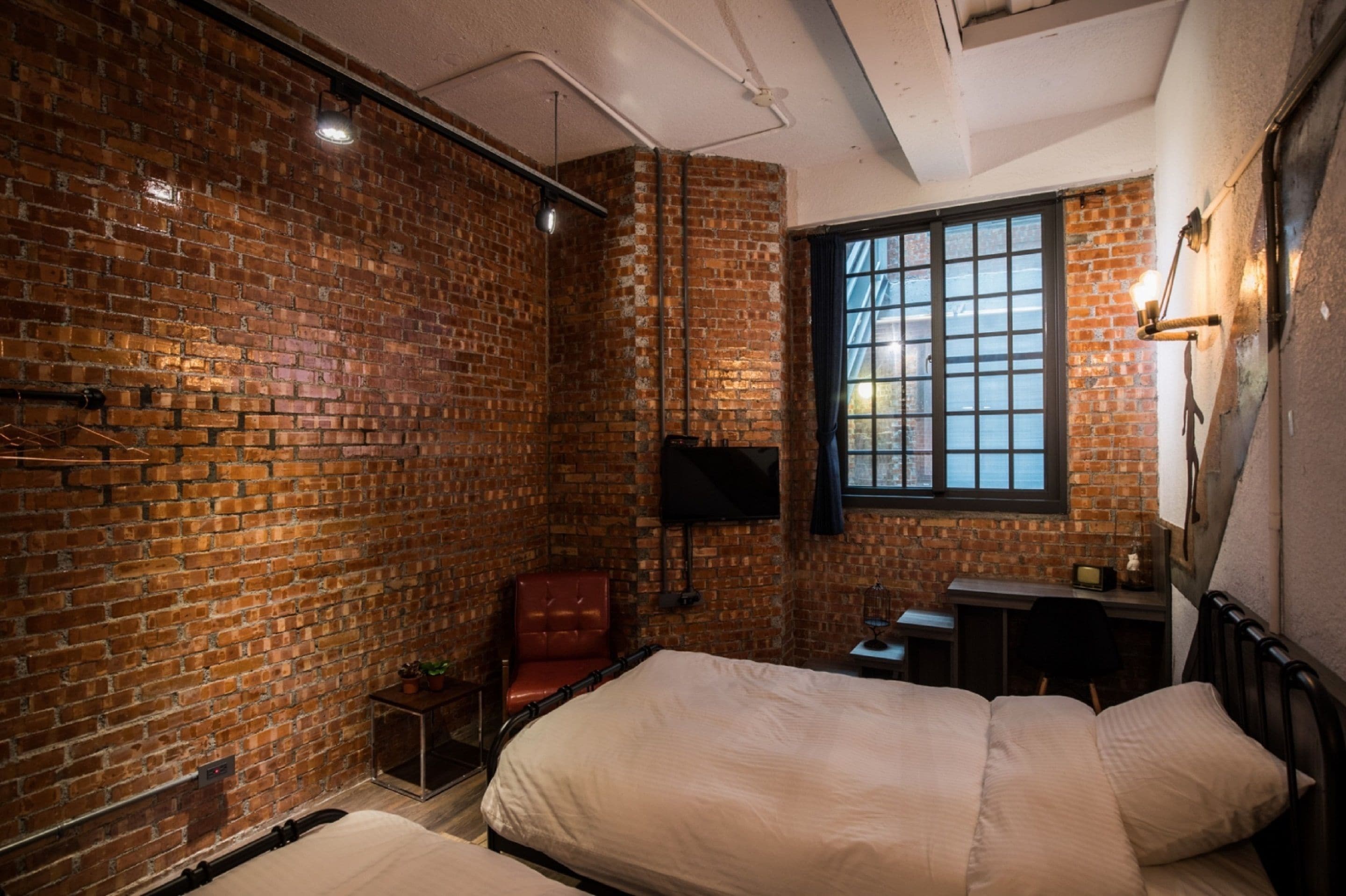A room with brick walls, a large window, a bed, a desk, and a chair.