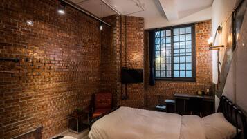 A room with brick walls, a large window, a bed, a desk, and a chair.