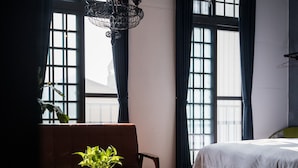 Ein Hotelzimmer mit einem Bett, einer Sitzecke mit Tisch und Stühlen, einer Zimmerpflanze und großen Fenstern mit Vorhängen.