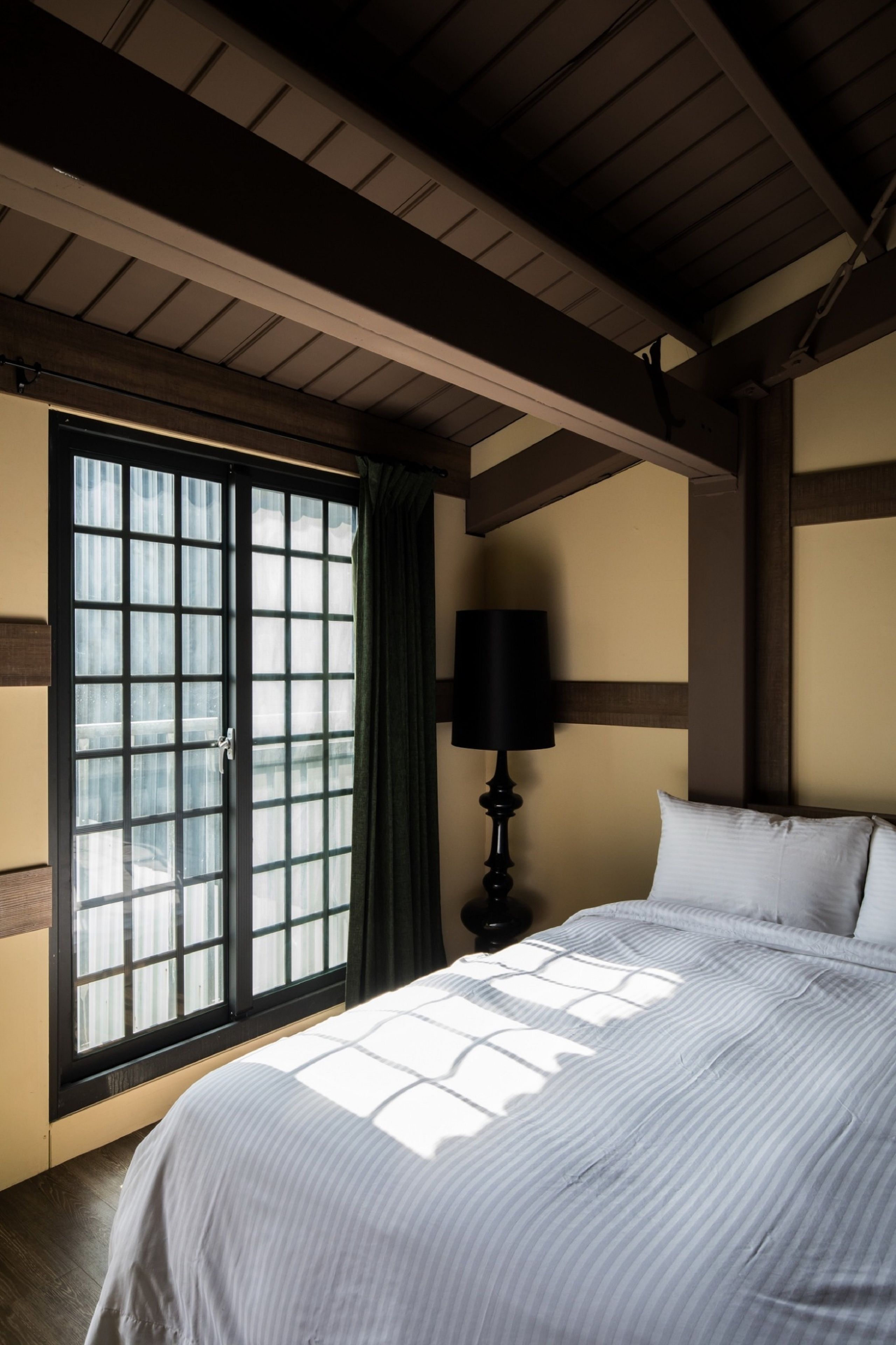 A bedroom with a large window, a bed with white bedding, a black lamp, and wooden beams on the ceiling.