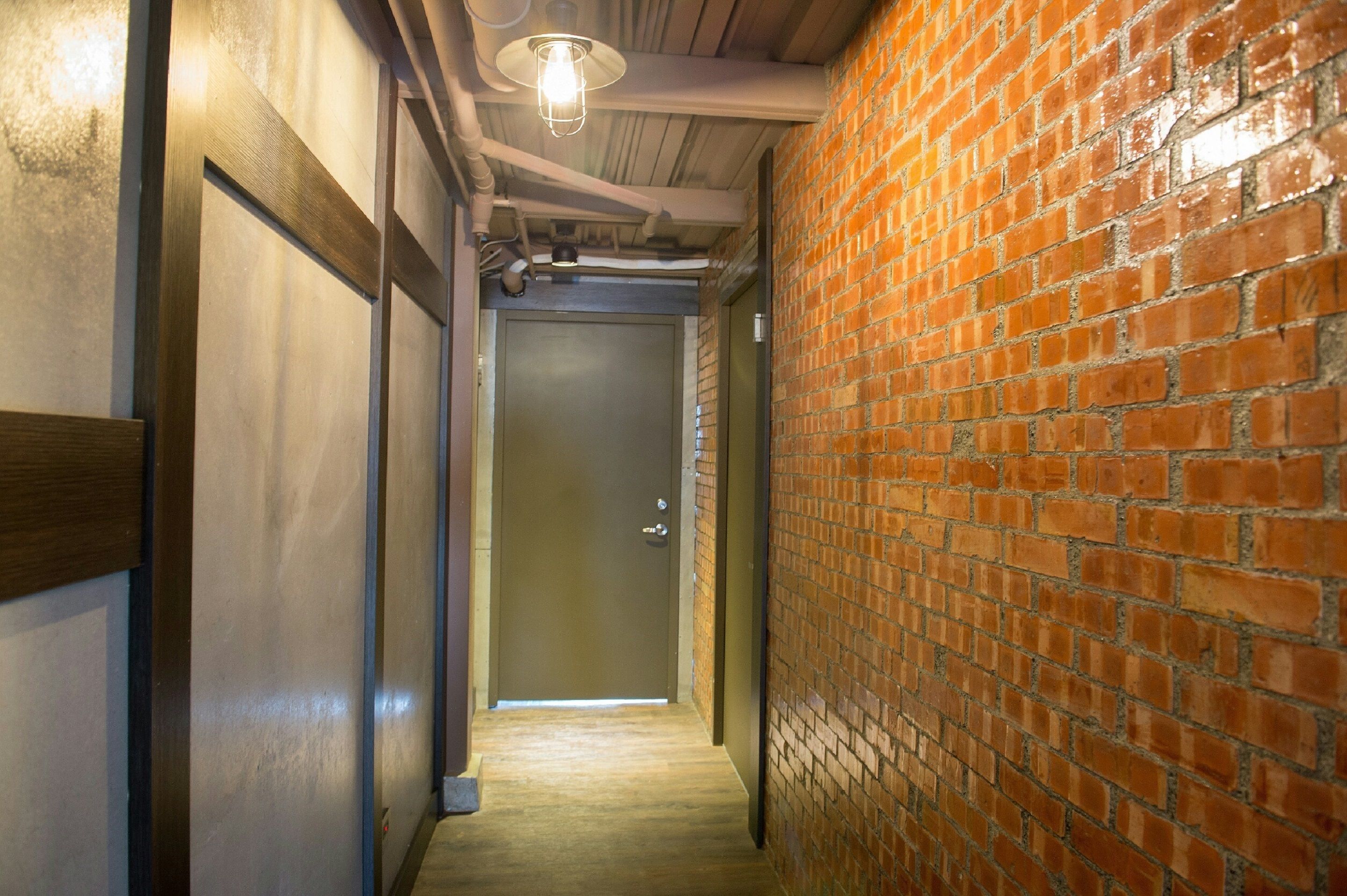 A narrow hallway with a wooden door at the end, a brick wall on the right, and a light fixture on the ceiling.
