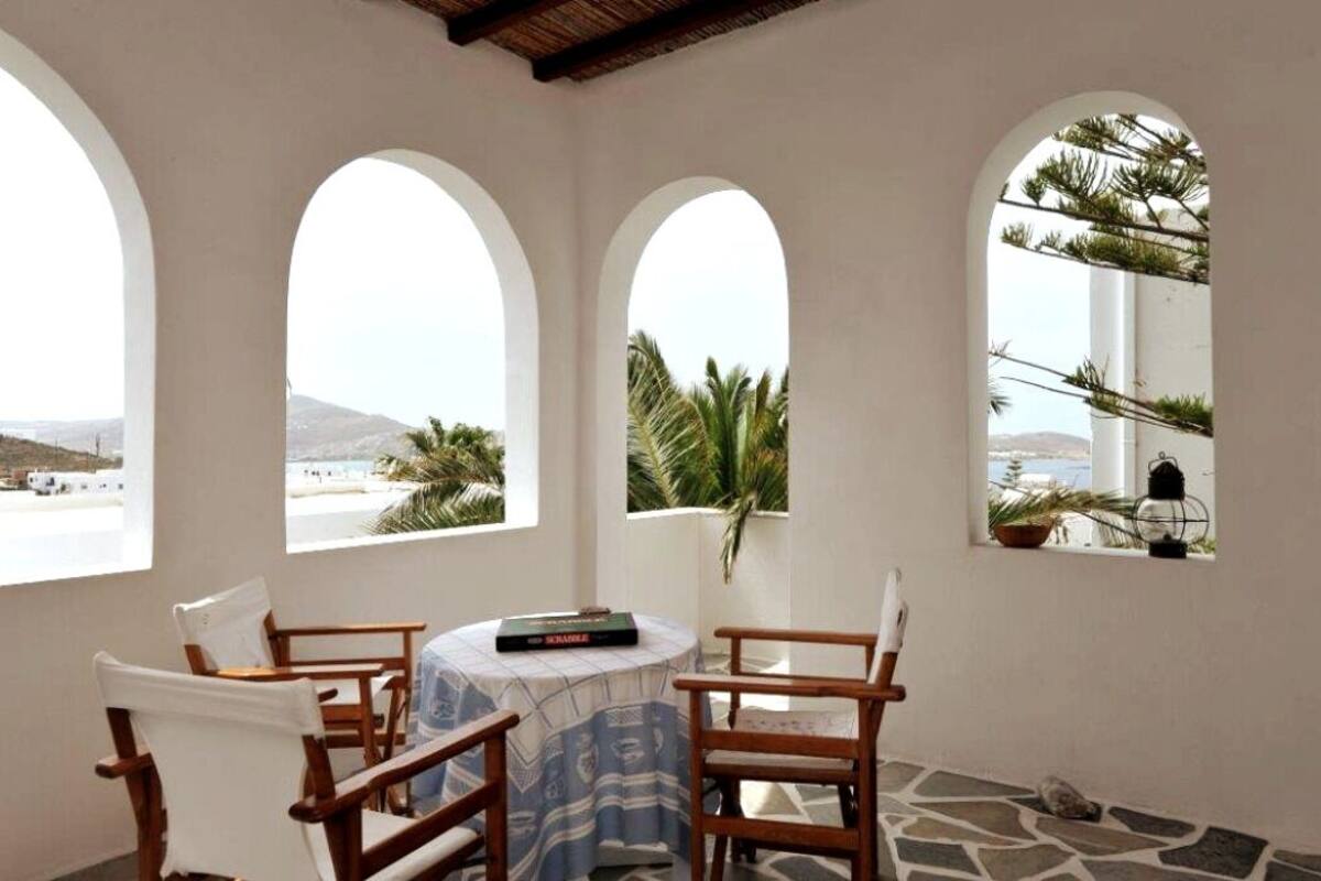 A room with three arched windows overlooking a beach, a round table with chairs, and a book on the table.