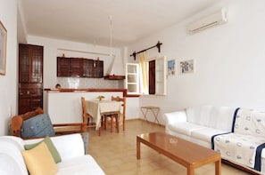 A living room with a sofa, a coffee table, and a dining area with a table and chairs. There is a kitchen area with cabinets and a window. - Rallis Apartments (Paros)