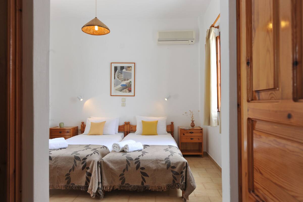 A double bed with a brown leaf-patterned bedspread, two bedside tables with lamps, a wall-mounted air conditioner, and a framed picture above the bed.