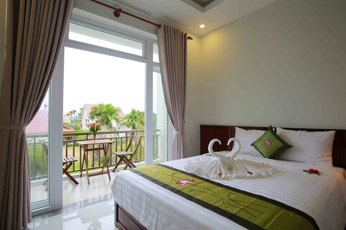 Habitación de hotel con balcón, una cama con colcha verde y una decoración de toallas con forma de cisne.