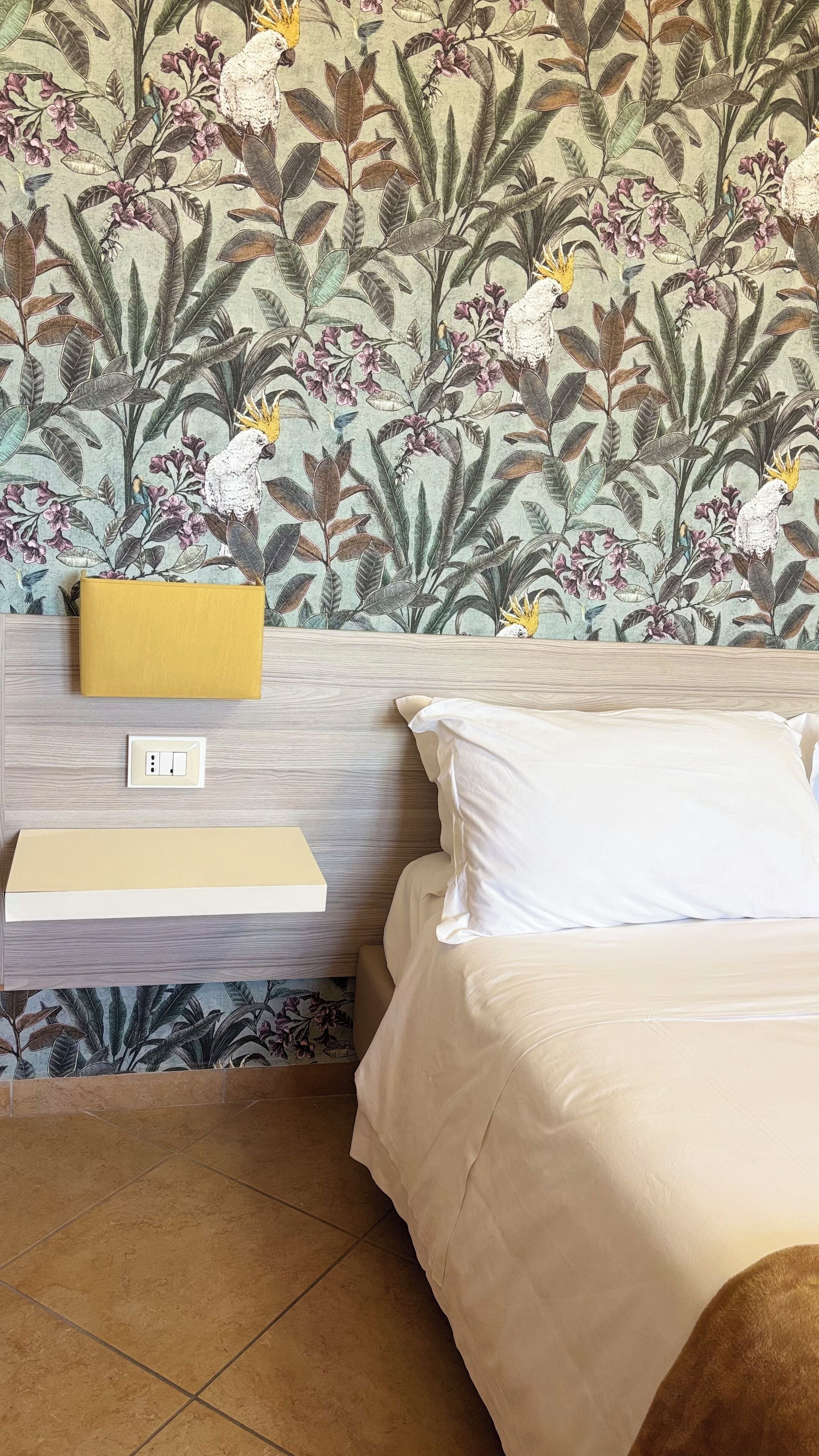 A bedroom with a bed, a bedside table, a lamp, and a wall with floral wallpaper.