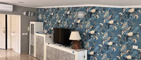 A modern living room with a tiled floor, a white entertainment unit, a flat-screen TV, a lamp, and a floral wallpaper.