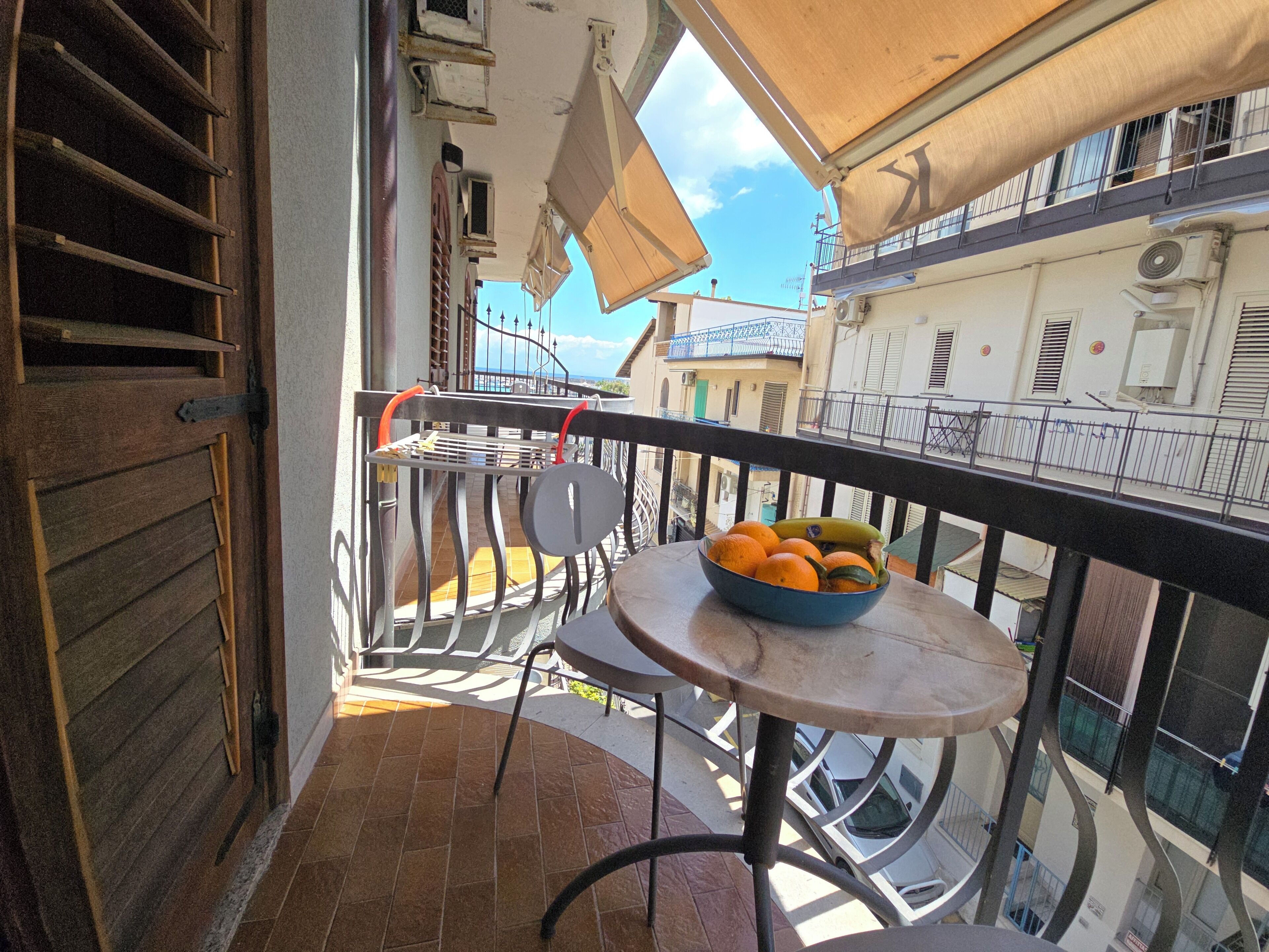 Un balcón con una mesa que sostiene naranjas y plátanos, una silla de metal y vistas de la ciudad.