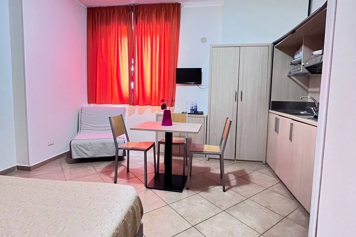 A compact hotel room with a bed, a dining table with chairs, a kitchenette, and a TV mounted on the wall.