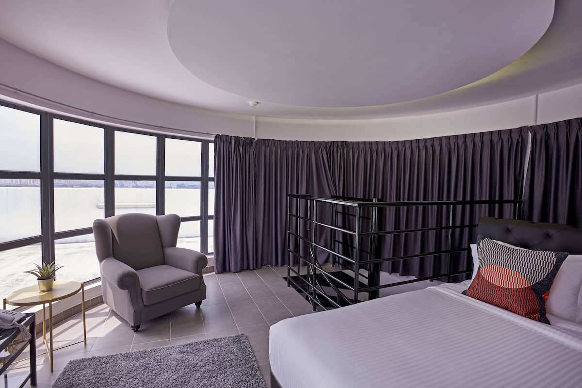 A modern hotel room with a large bed, a seating area with a chair, a small table, and a view of the water.