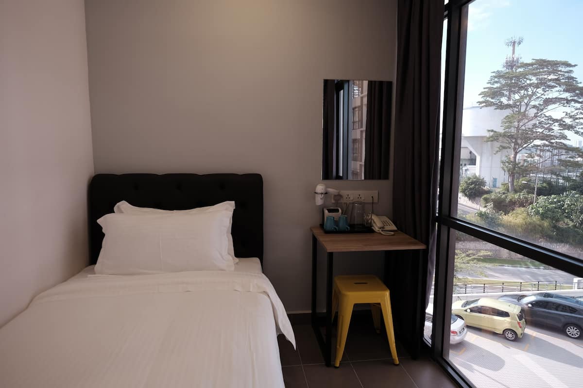 A hotel room with a bed, a small table with a telephone and a coffee maker, a stool, and a window with a view of parked cars and trees.