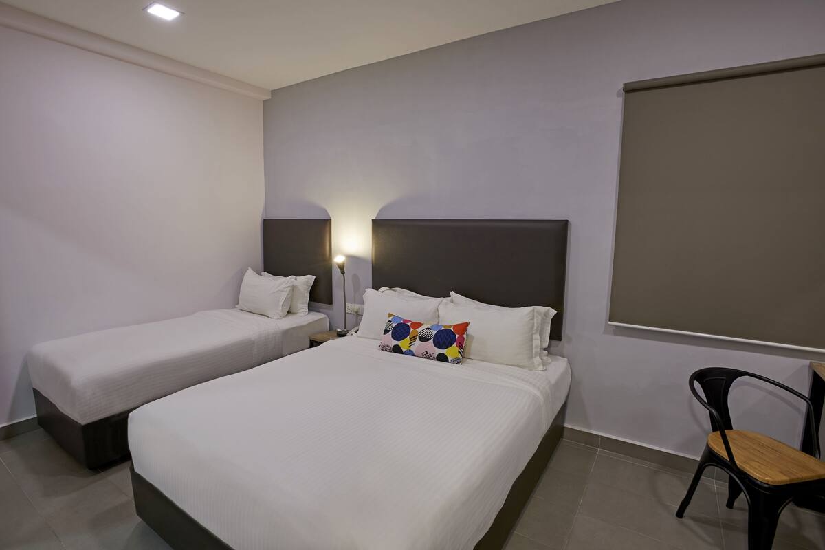 A hotel room with two single beds, a large bed, a bedside table with a lamp, a chair, and a large window.