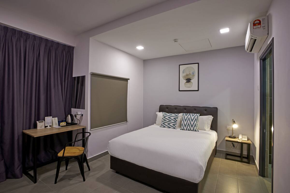 A hotel room with a bed, a desk, a chair, a bedside table with a lamp, an air conditioner, and a window with curtains.