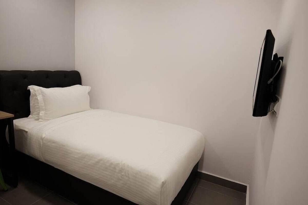 A small, single-bed room with a dark-colored headboard, a white bedspread, and a flat-screen TV mounted on the wall.
