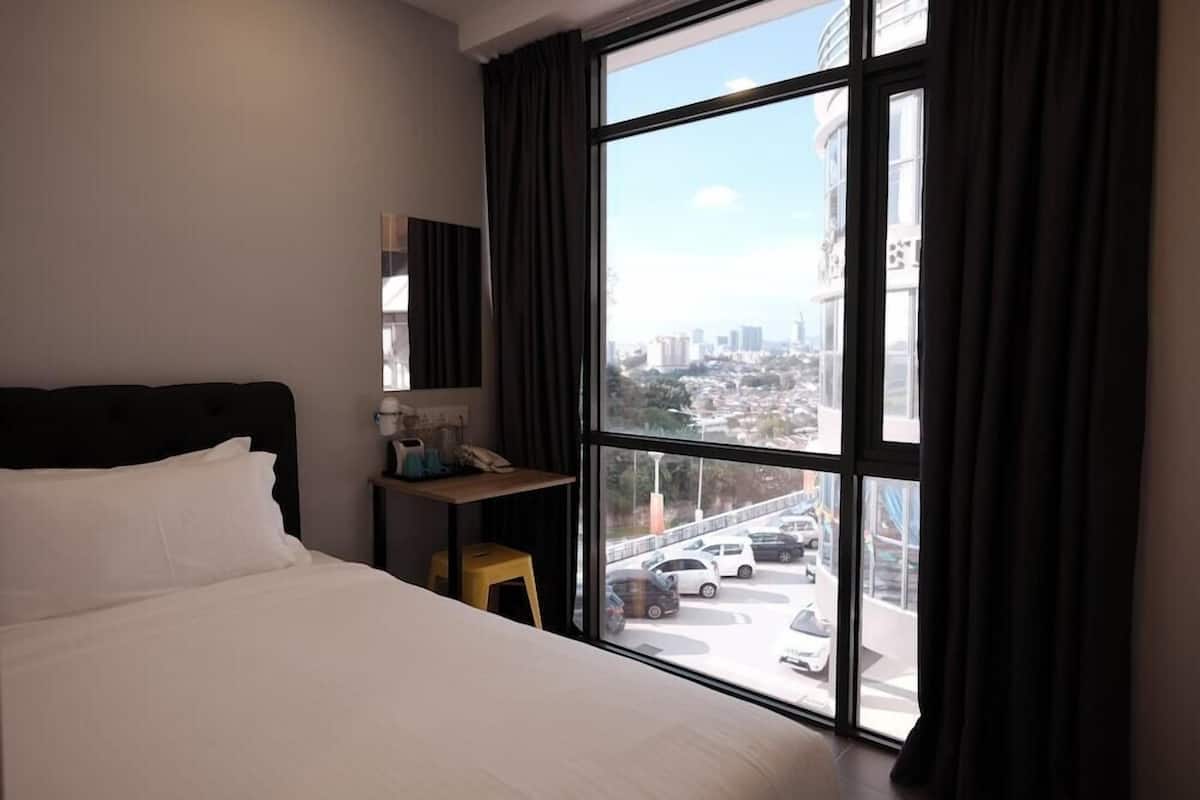 A hotel room with a large window, a bed, a desk with a chair, and a small table with a lamp.