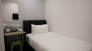 Habitación de hotel pequeña y ordenada con una cama individual, una mesita de noche con lámpara, un espejo y un taburete verde.