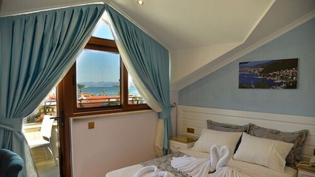 A bedroom with a bed, a nightstand, a window with curtains, a painting on the wall, and a view of the sea. - Kayi Hotel (Fethiye)