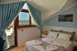 A bedroom with a bed, a nightstand, a window with curtains, a painting on the wall, and a view of the sea.