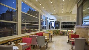 A restaurant with a modern interior, featuring a mix of red and grey upholstered chairs, a variety of table shapes, and large windows offering views of the cityscape at night. - Kayi Hotel (Fethiye)