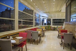 Een restaurant met een modern interieur, met rode en grijze bekleden stoelen, verschillende tafelvormen en grote ramen met uitzicht op de stad 's nachts.