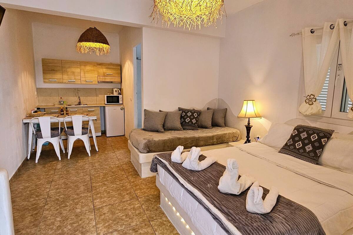 A bedroom with a bed, bedside table, lamp, and towels. There is a kitchenette with a microwave and a dining area with chairs and a table.